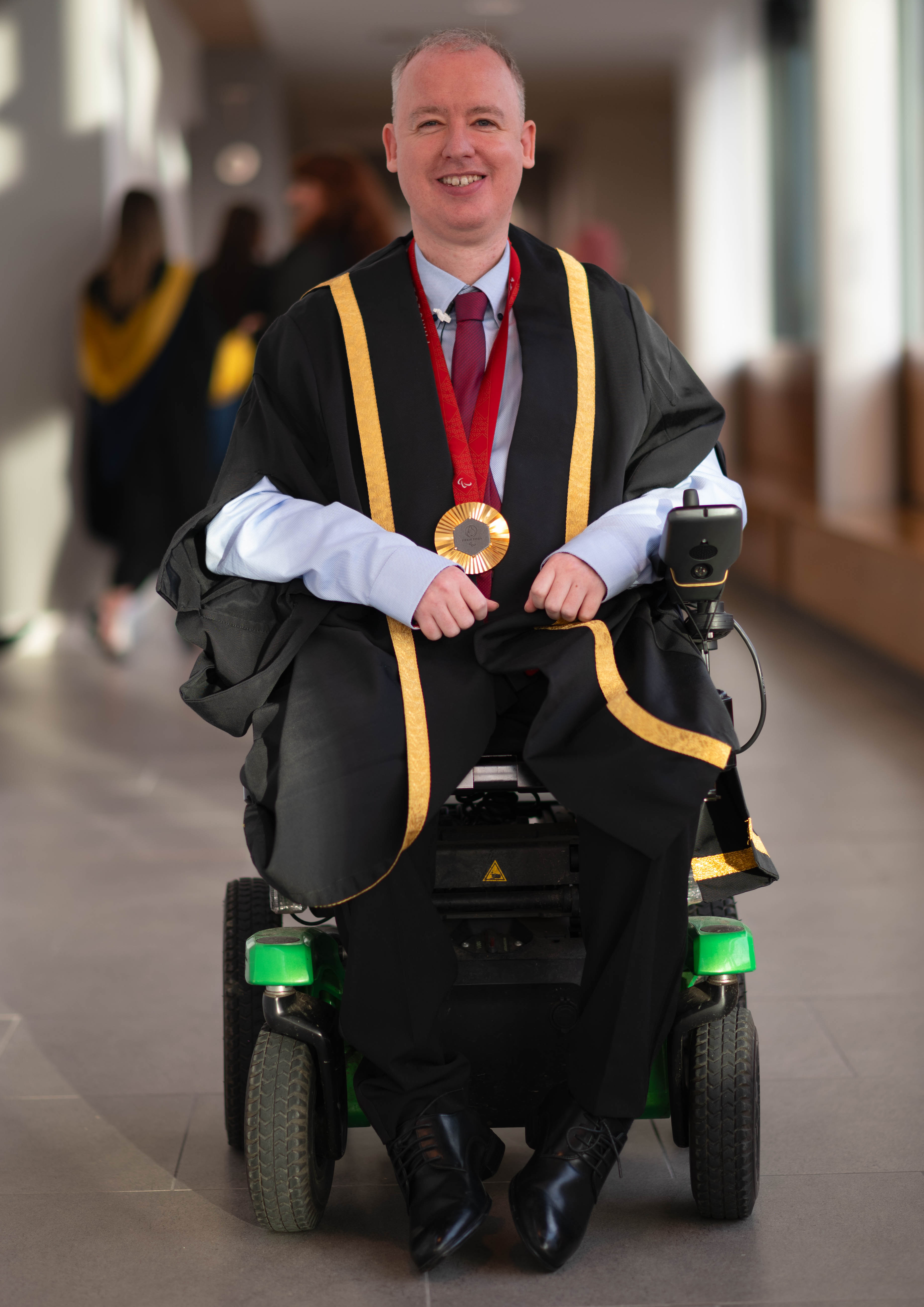 Stephen McGuire, a Paralympic gold medallist, wearing formal graduation attire with gold trim and a medal around his neck, seated in a wheelchair. He is smiling and positioned in a well-lit hallway, with blurred figures of graduates in the background.